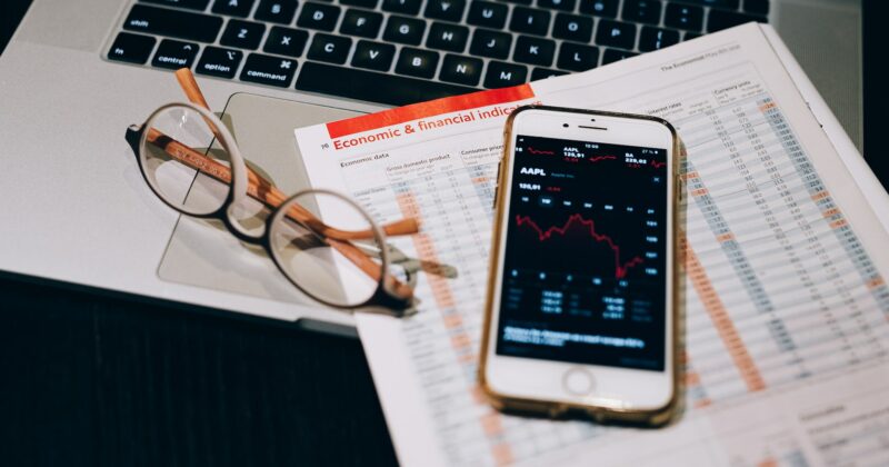 A laptop displaying one of the 20 best finance websites, alongside a cell phone and glasses, rests on a desk.